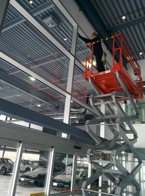 A man on a scissor lift to clean tall windows in a car dealership.